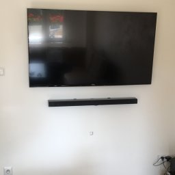 Remonty.GZ - Telewizor z soundbarem zamontowany na ścianie w jasnym pokoju, widoczne gniazdka elektryczne i fragment podłogi.