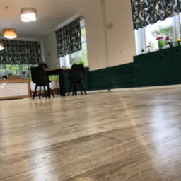 Jasne panele podłogowe z widoczną strukturą drewna, w tle fragment odnowionego wnętrza z ciemnymi krzesłami i zasłonami w roślinne wzory.