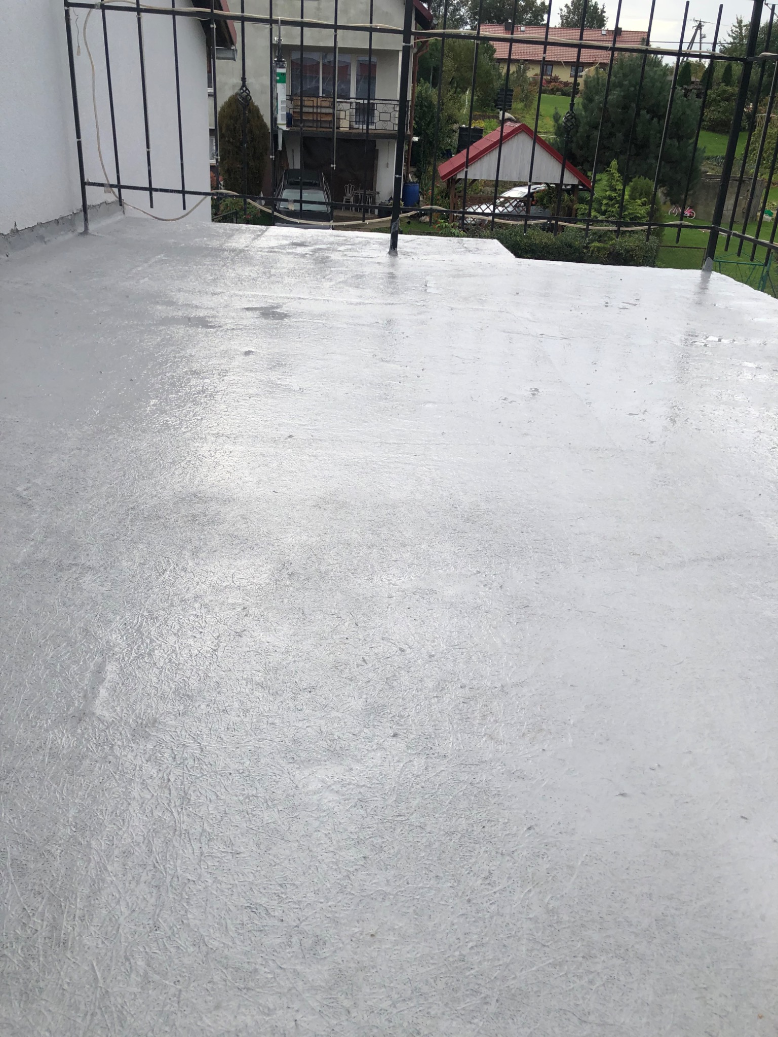 Szara, świeżo wylana posadzka epoksydowa na balkonie, widok z góry na teksturę powierzchni i metalową barierkę, w tle zabudowa jednorodzinna i zieleń.