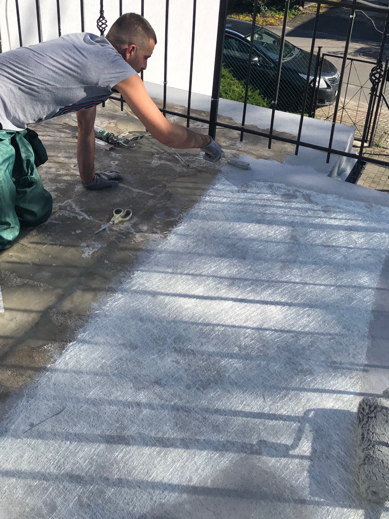 Pracownik w szarych rękawicach nakłada żywicę na włókno szklane na balkonie, widoczne nożyczki, wałek malarski i zielone spodnie robocze.