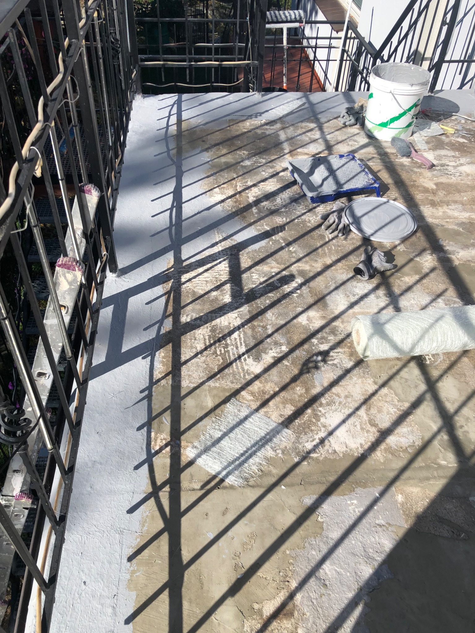 Balkon w trakcie malowania: widoczne narzędzia malarskie, wiadro z farbą i częściowo pomalowana powierzchnia podłogi z widocznymi smugami i nierównościami, rzucane cienie krat balustrady.