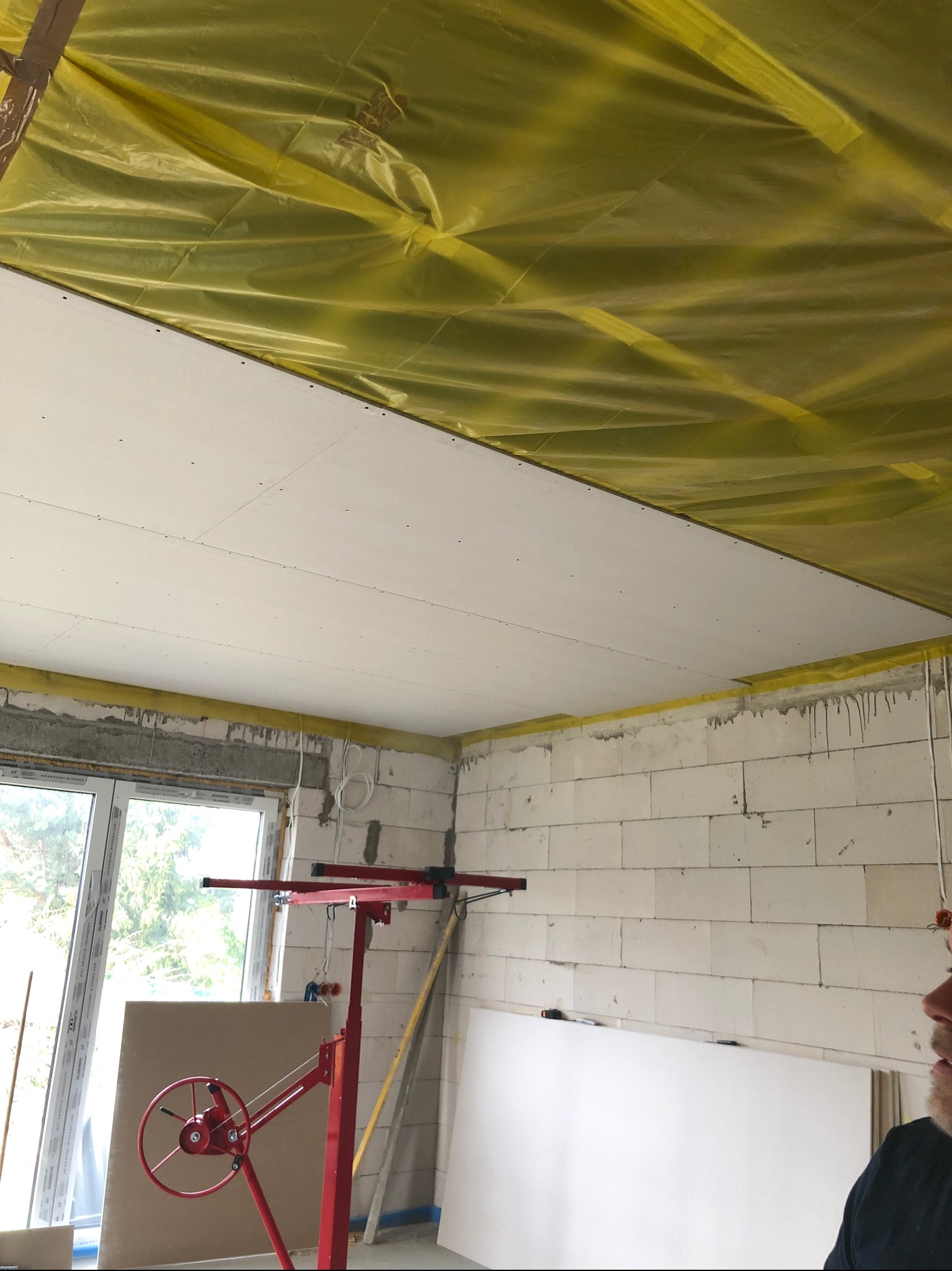 Montaż płyty gipsowo-kartonowej na suficie, widoczna podpora montażowa, folia ochronna na stropie, surowy stan ścian z betonu komórkowego, okno z widokiem na zieleń.