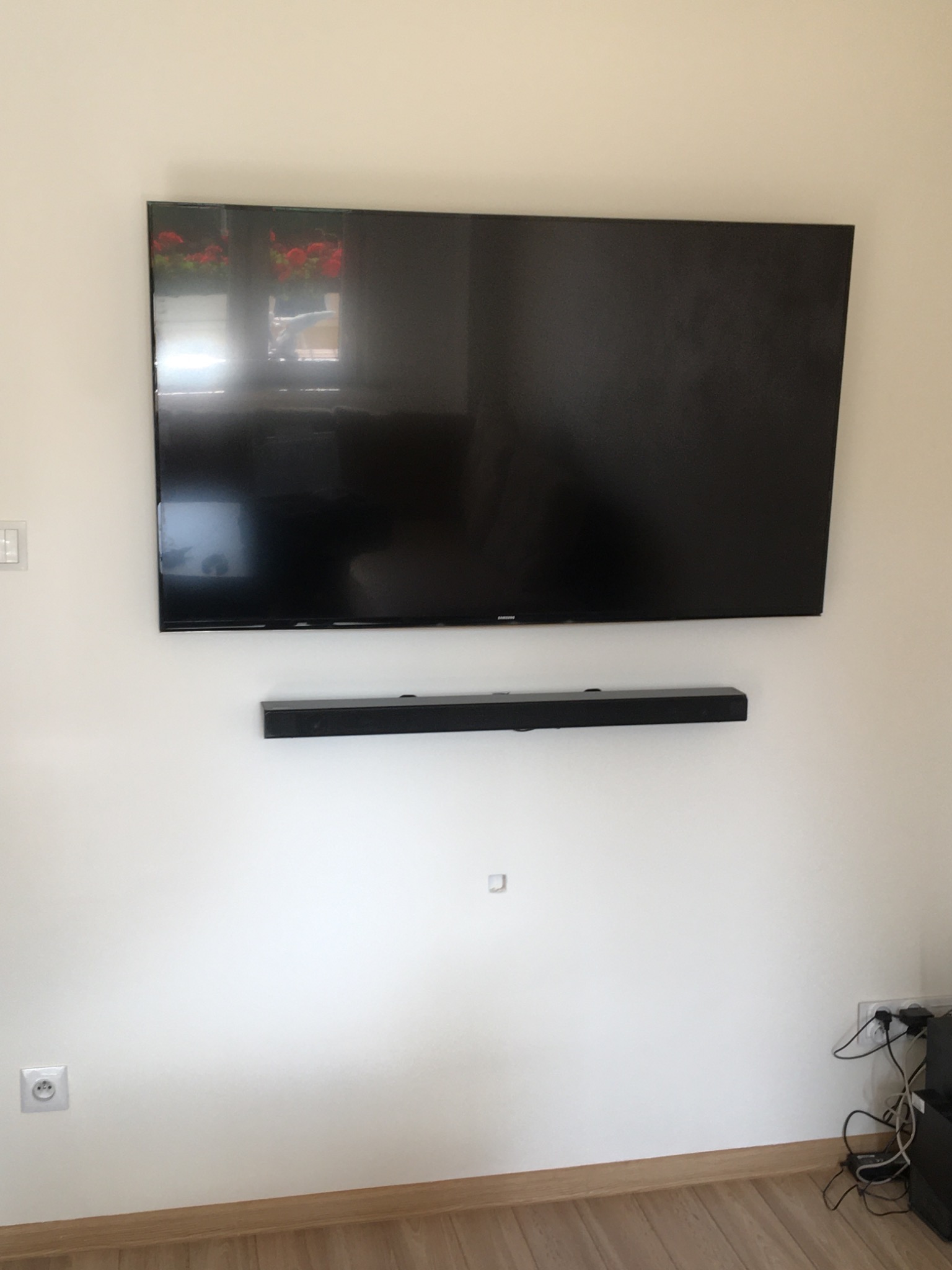 Telewizor z soundbarem zamontowany na ścianie w jasnym pokoju, widoczne gniazdka elektryczne i fragment podłogi.