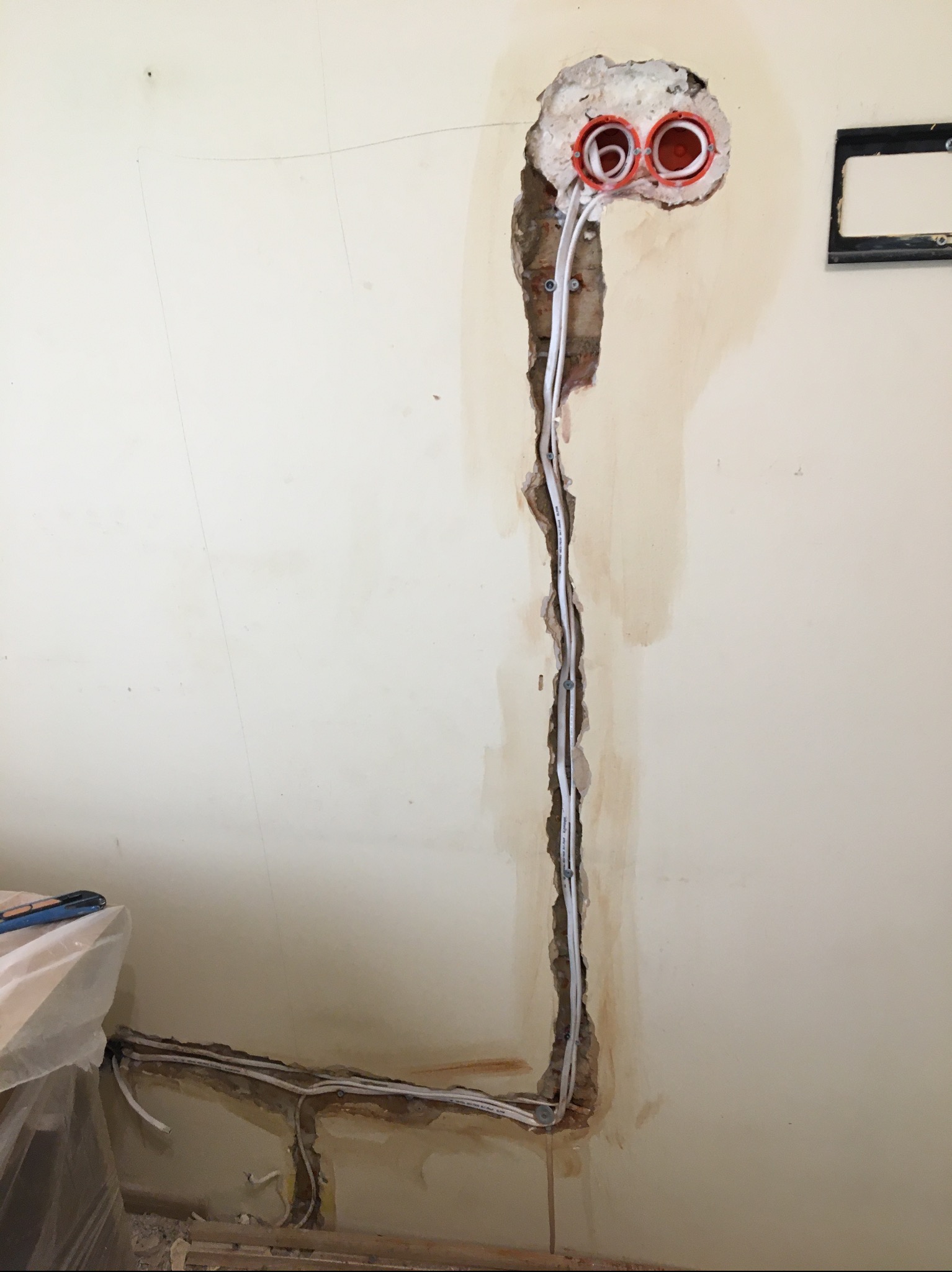 Ściana z wyciętym bruzdą pod kable elektryczne, widoczne pomarańczowe puszki instalacyjne i białe przewody.