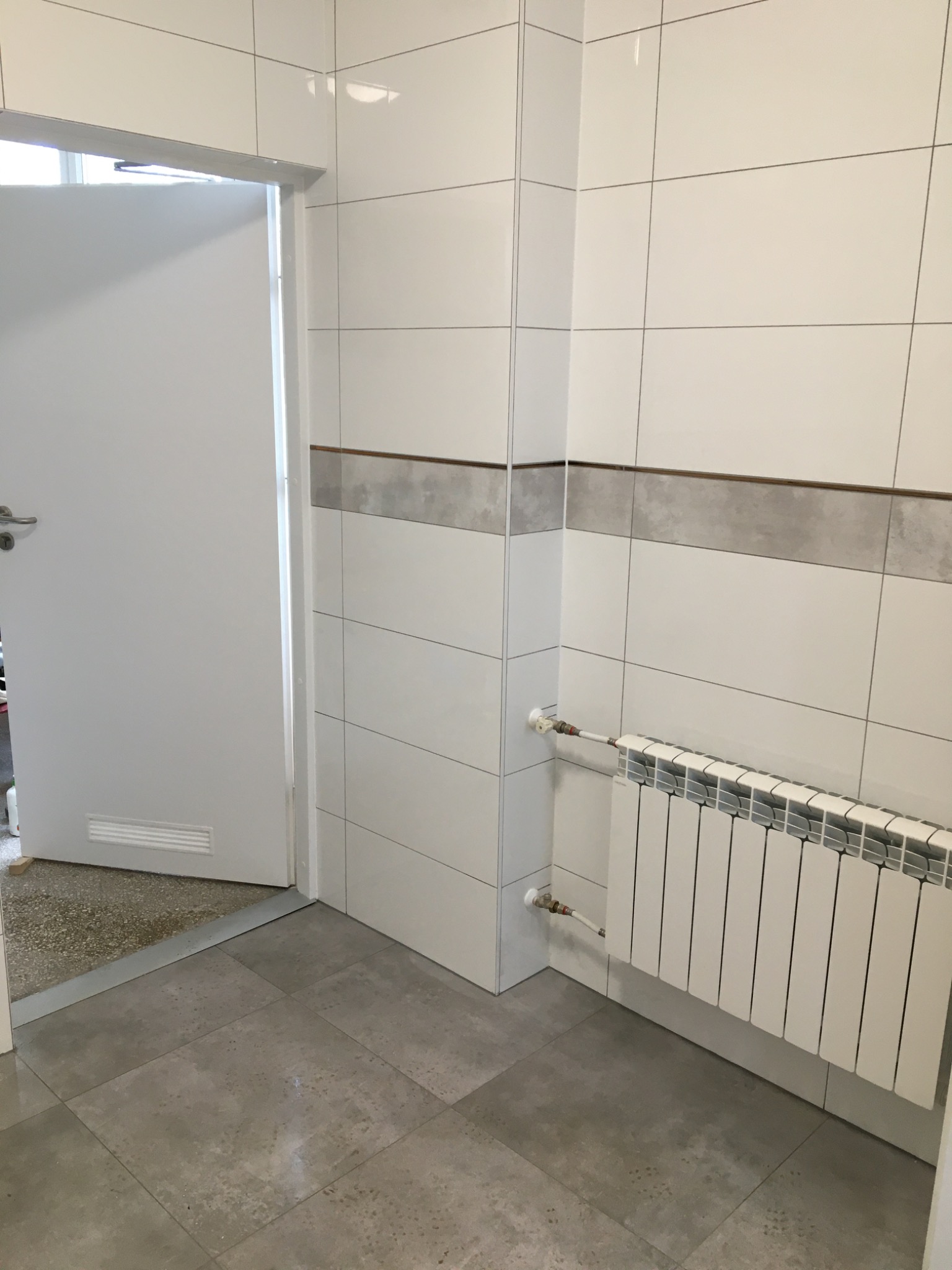 Wykończona łazienka z białymi płytkami, szarym pasem dekoracyjnym, grzejnikiem i otwartymi białymi drzwiami.