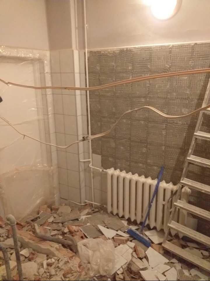 Łazienka w trakcie demontażu płytek, widoczne gruz, grzejnik, drabina i odsłonięta ściana pod przyszłe płytki.