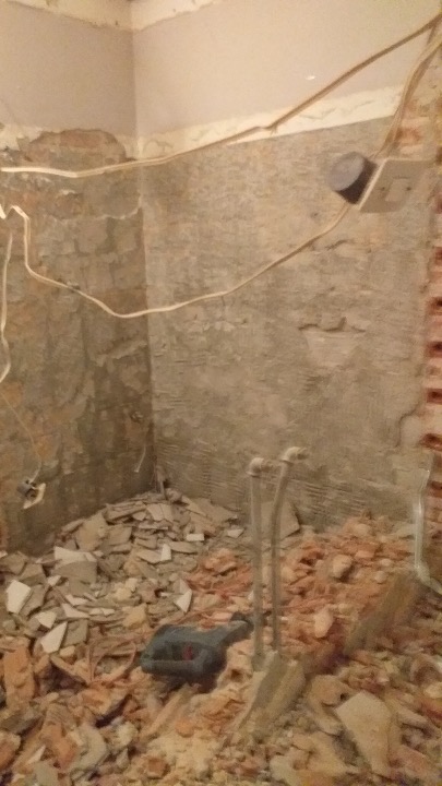 Łazienka w trakcie demontażu płytek, widoczne gruz, wystające rury, przewody elektryczne i narzędzie udarowe na podłodze.