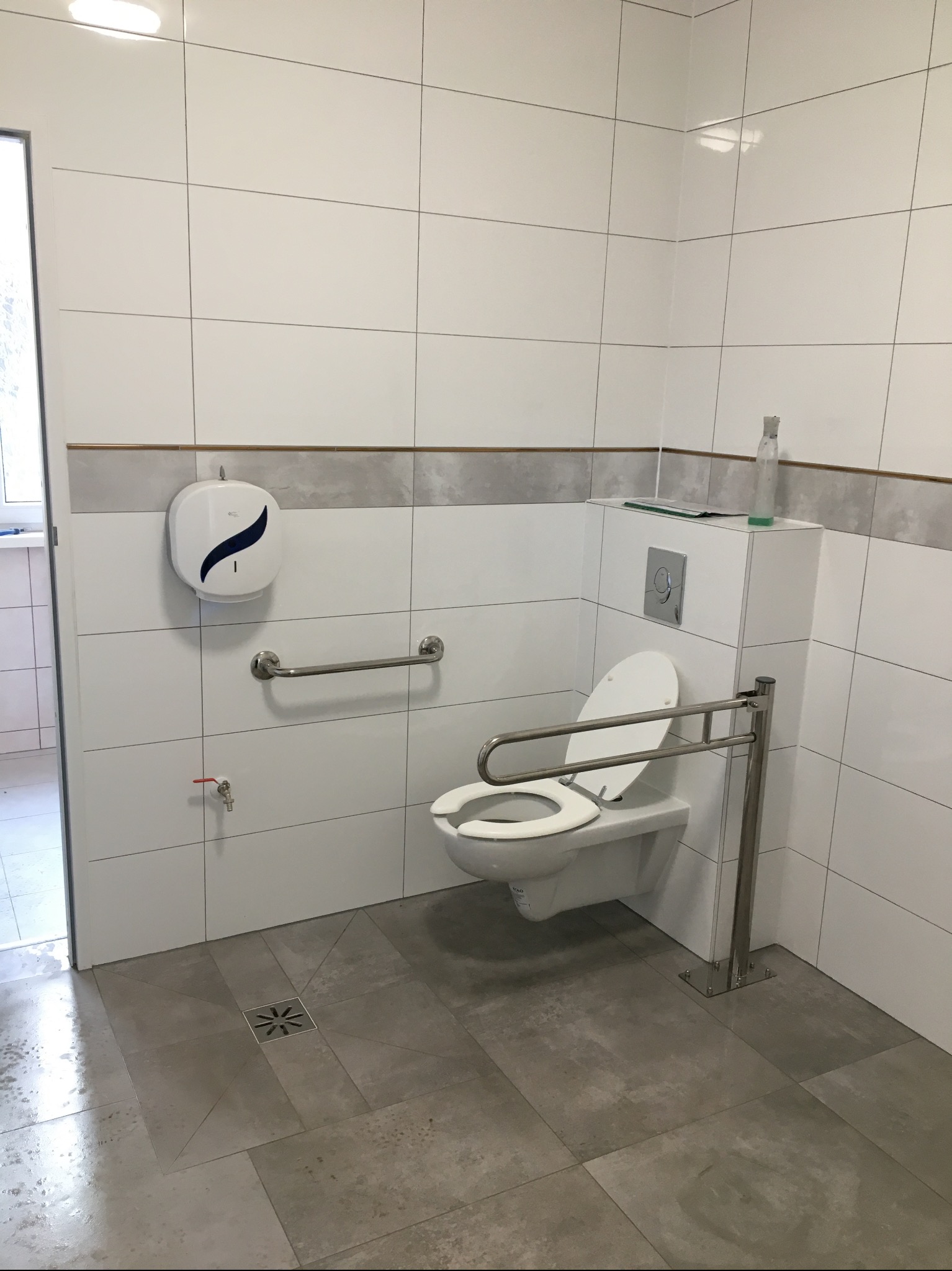 Wykończona łazienka z toaletą dla niepełnosprawnych, białe płytki z szarym pasem dekoracyjnym, uchwyty ze stali nierdzewnej, podłoga z szarych płytek, widoczny dozownik mydła i butelka z płynem.