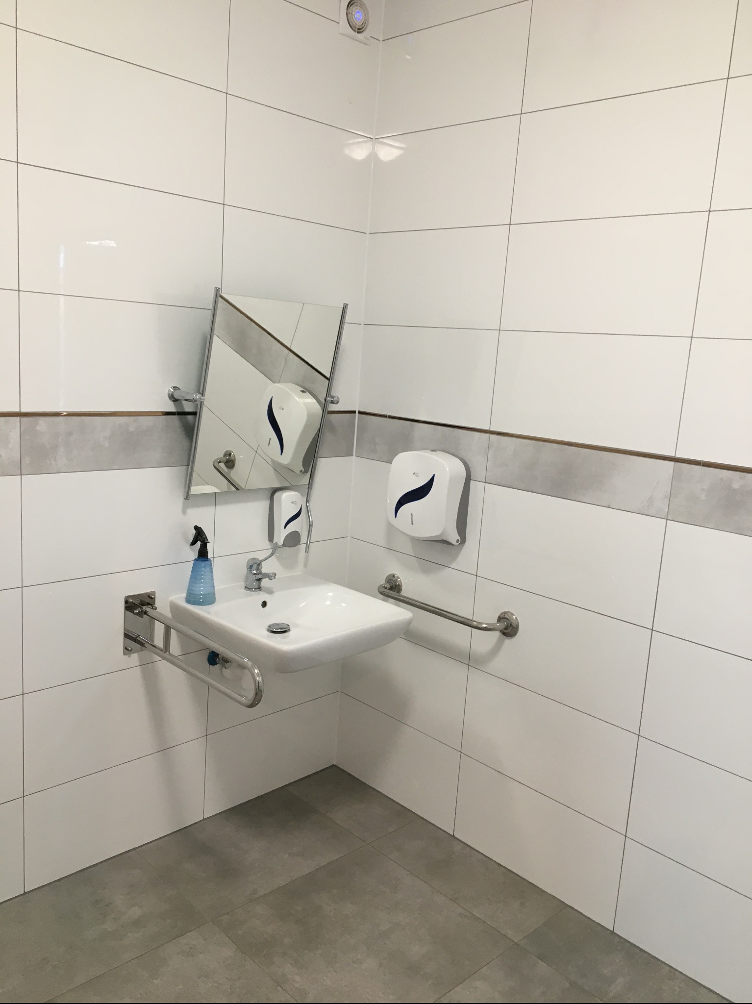 Nowoczesna łazienka z białą umywalką dla niepełnosprawnych, chromowanymi uchwytami, lustrem i dozownikiem mydła, wykończona białymi płytkami z szarym pasem dekoracyjnym i szarą podłogą z płytek.