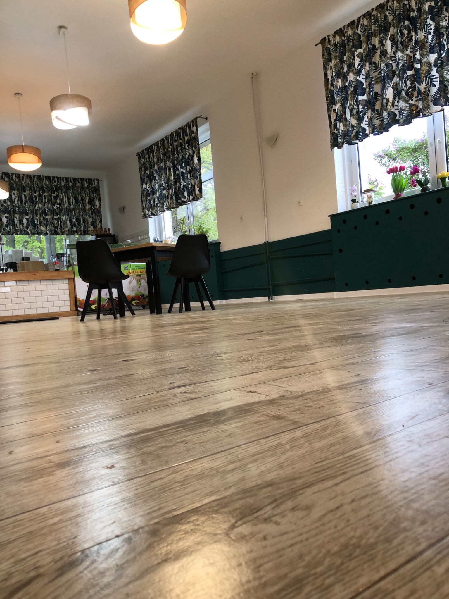 Jasne panele podłogowe z widoczną strukturą drewna, w tle fragment odnowionego wnętrza z ciemnymi krzesłami i zasłonami w roślinne wzory.