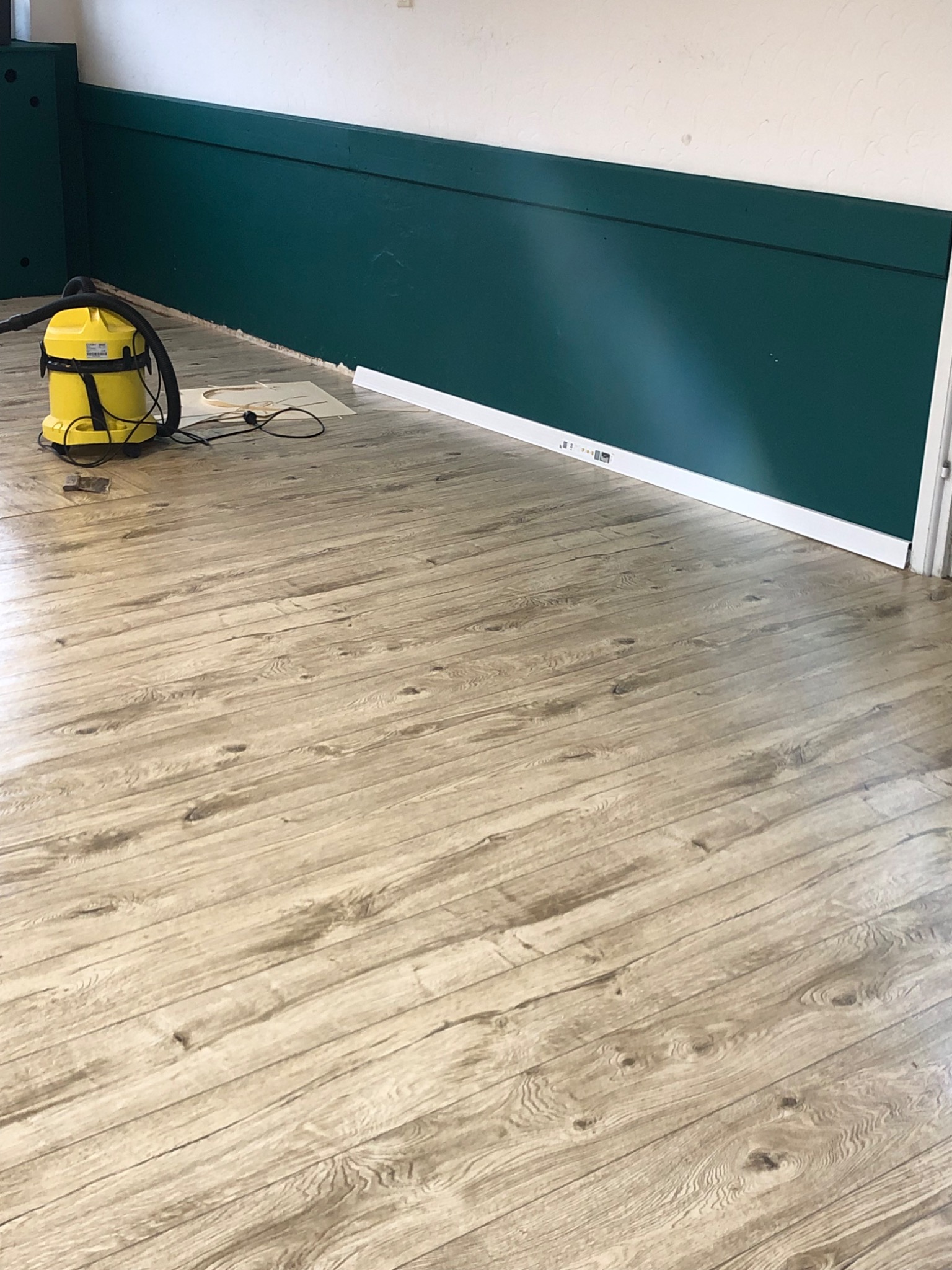 Świeżo ułożone panele podłogowe o wzorze jasnego drewna, z widocznym żółtym odkurzaczem przemysłowym i fragmentami listew przypodłogowych przy ścianie pomalowanej na ciemnozielono.