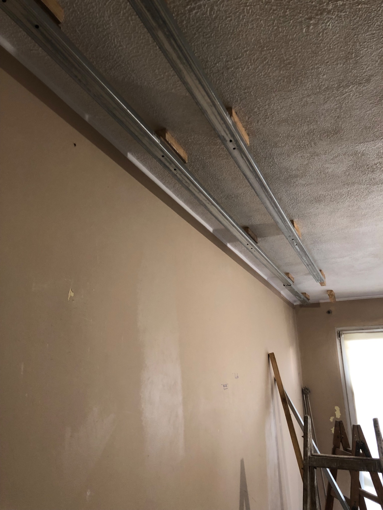 Montaż metalowych profili sufitowych na drewnianych wspornikach, widoczna ściana w kolorze beżowym i drabina oparta o ścianę