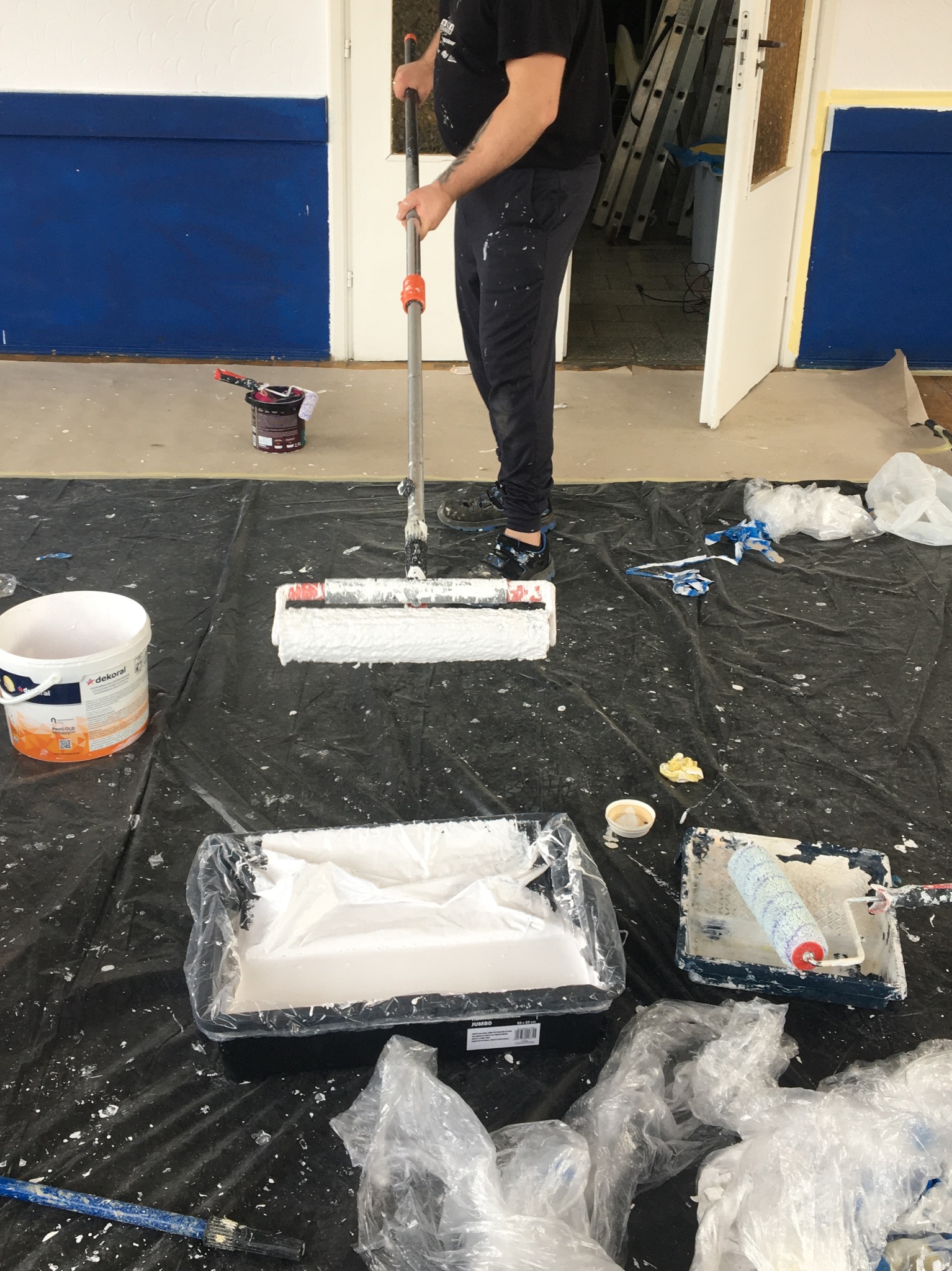 Mężczyzna maluje ścianę wałkiem na długim kiju, widoczne kuwety z farbą, wiadro z farbą Dekoral i folia ochronna na podłodze.