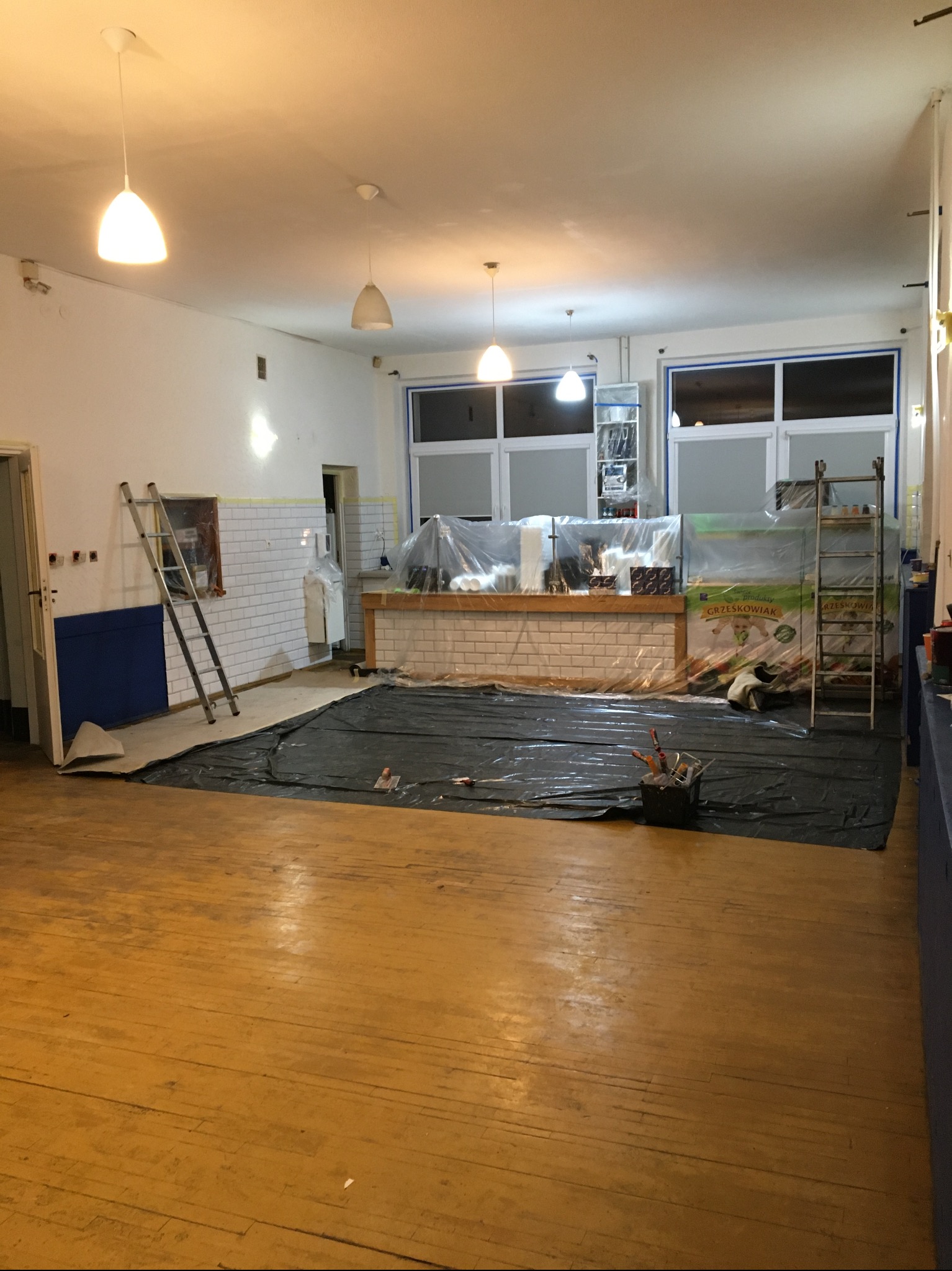 Wnętrze w trakcie przygotowań do malowania: podłoga zabezpieczona folią, ściany z częściowo nałożonym kolorem, drabina oparta o ścianę, widoczne wiadro z narzędziami malarskimi.