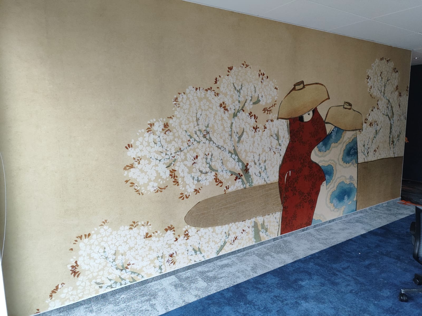 Beżowa ściana z ręcznie malowanym, japońskim motywem kwitnącej wiśni i postaci w tradycyjnych strojach. Dekoracyjne wykończenie wnętrza z artystycznym akcentem.