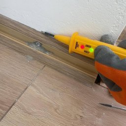 LIVANS SPÓŁKA Z OGRANICZONĄ ODPOWIEDZIALNOŚCIĄ - Żółty tester napięcia Phaser 777 w dłoni w rękawicy, sprawdzający przewody przy listwie przypodłogowej na tle drewnianej podłogi i białej ściany. Kontrolka napięcia świeci.