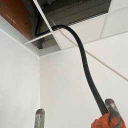 LIVANS SPÓŁKA Z OGRANICZONĄ ODPOWIEDZIALNOŚCIĄ - Czarny peszel instalacji elektrycznej prowadzony w przestrzeni sufitu podwieszanego, widoczny fragment drabiny i rękawicy roboczej.