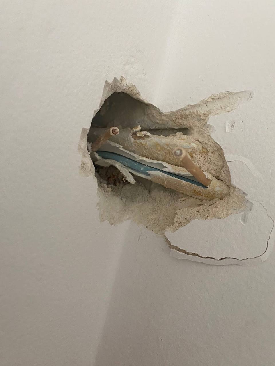 Uszkodzone przewody elektryczne wystające z dziury w ścianie, z widoczną niebieską izolacją i fragmentami tynku. Ujęcie z bliska, ukazujące detale uszkodzenia.