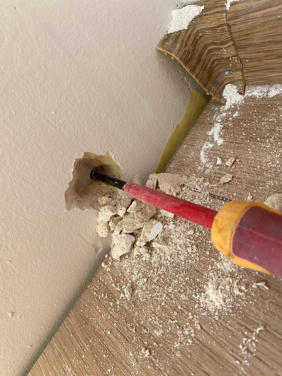 Uszkodzona ściana i podłoga po naprawie przewodu elektrycznego. Widoczny fragment przewodu i narzędzie z czerwoną rączką. Drobne uszkodzenia wokół otworu.