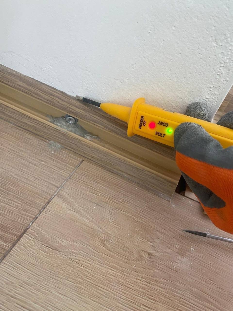 Żółty tester napięcia Phaser 777 w dłoni w rękawicy, sprawdzający przewody przy listwie przypodłogowej na tle drewnianej podłogi i białej ściany. Kontrolka napięcia świeci.