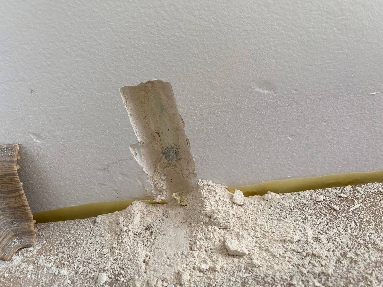 Uszkodzony, wystający ze ściany przewód w białej osłonie, otoczony gruzem i pyłem. Widoczne ślady naprawy. Fragment drewnianej listwy przypodłogowej po lewej.