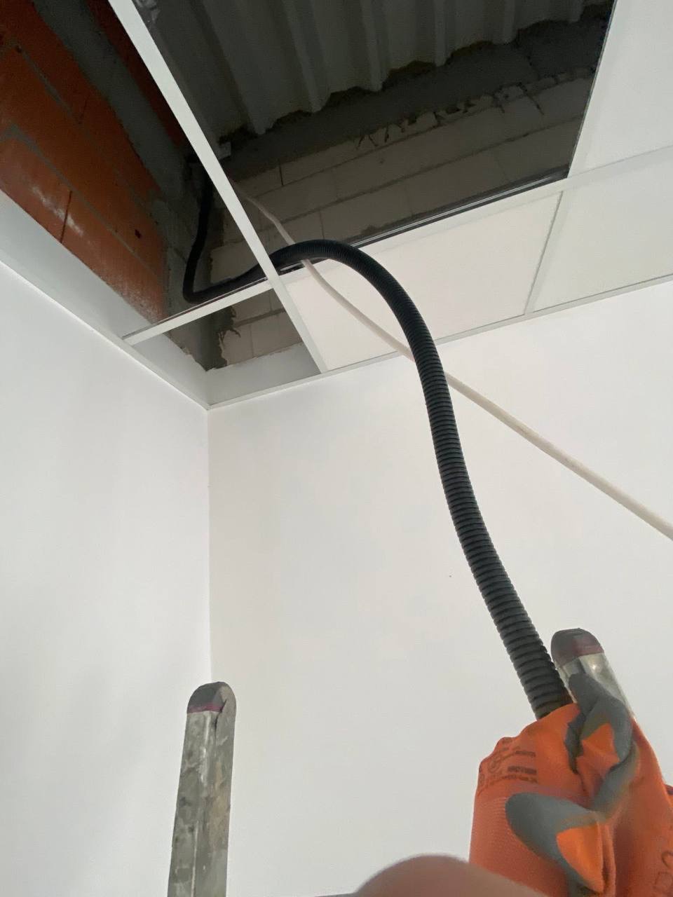 Czarny peszel instalacji elektrycznej prowadzony w przestrzeni sufitu podwieszanego, widoczny fragment drabiny i rękawicy roboczej.