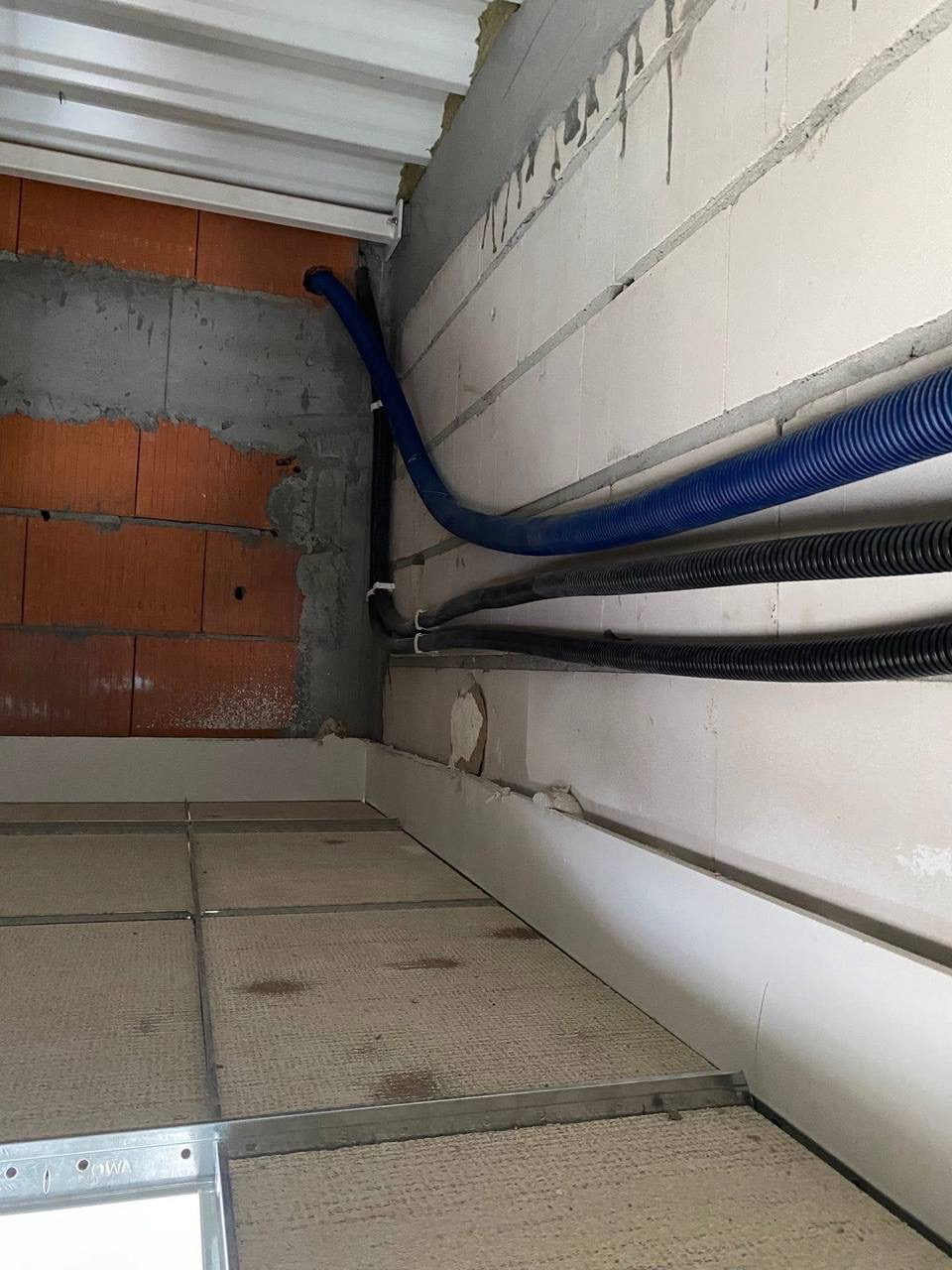 Niebieskie i czarne rury karbowane prowadzone wzdłuż ściany z białych bloczków, z widocznym fragmentem instalacji pod sufitem podwieszanym.