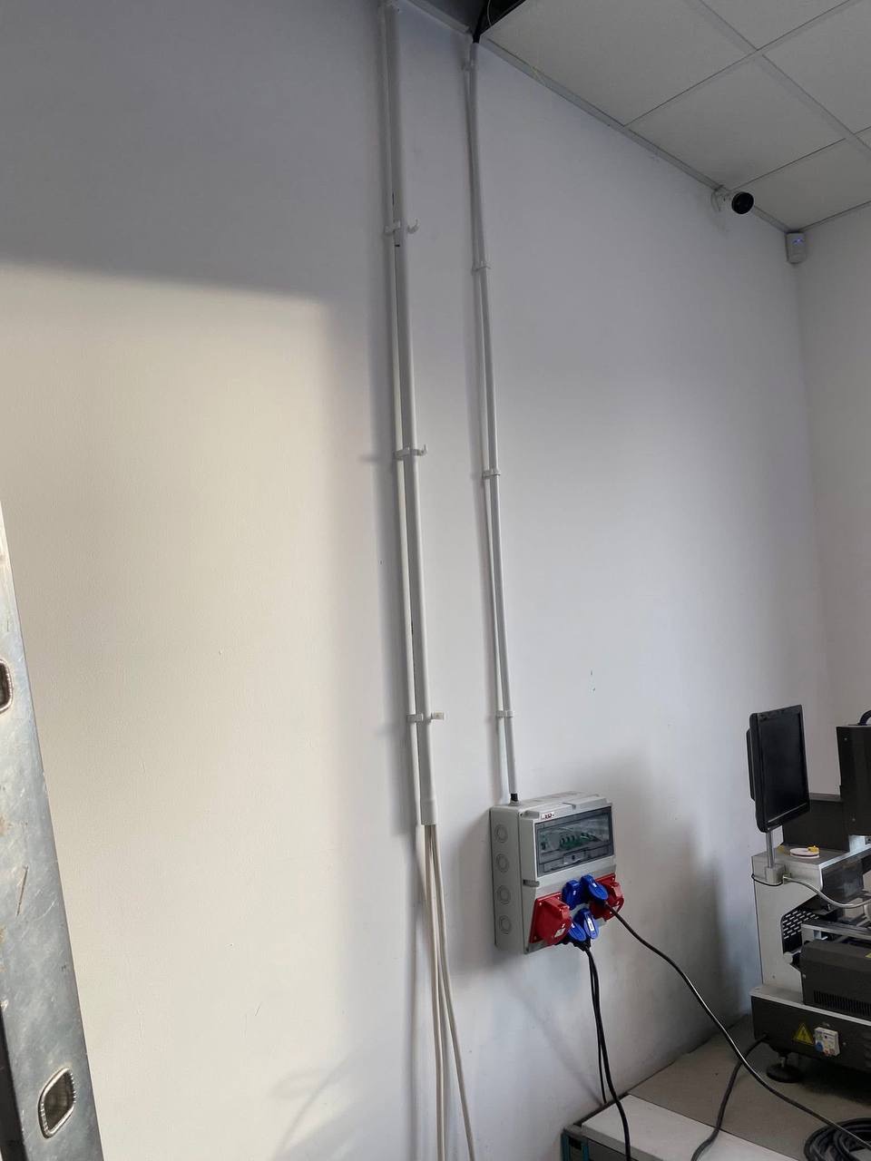 Szafa elektryczna z podłączonymi przewodami, zamontowana na białej ścianie. Widoczne rury instalacyjne i fragment drabiny. W tle sprzęt elektroniczny i kamera przemysłowa.