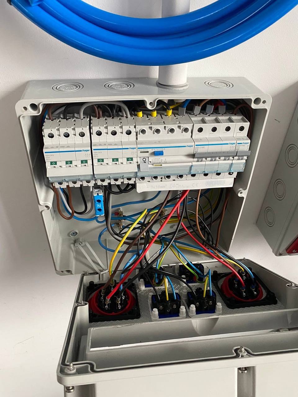 Otwarta szafa elektryczna z widocznymi bezpiecznikami, przewodami i elementami przyłączeniowymi, zamontowana na białej ścianie, z niebieską rurą instalacyjną u góry.