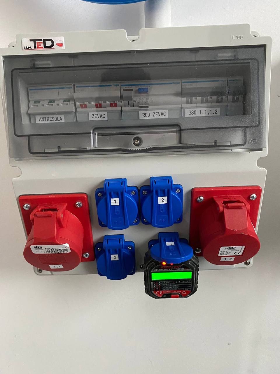 Szafa elektryczna z bezpiecznikami, gniazdami przemysłowymi i testerem gniazd, pokazująca wykonane podłączenie zasilania do maszyny. Widoczne oznaczenia i etykiety.