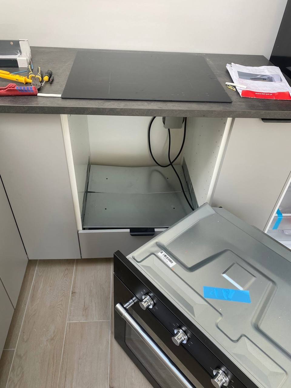 Płyta indukcyjna na blacie kuchennym, pod spodem widoczne kable. Obok leżą narzędzia i dokumentacja. Piekarnik czeka na montaż. Ujęcie z góry, neutralne oświetlenie.