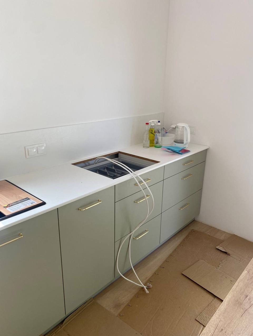 Podłączenie płyty indukcyjnej w kuchni: biały blat, oliwkowe szafki z złotymi uchwytami, widoczne przewody elektryczne i karton zabezpieczający podłogę.