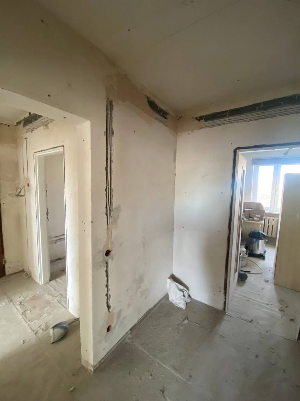 Wnętrze w trakcie remontu: bruzdy pod kable elektryczne w ścianie, widoczne przewody, przygotowanie do tynkowania. W tle odkurzacz przemysłowy i okno.