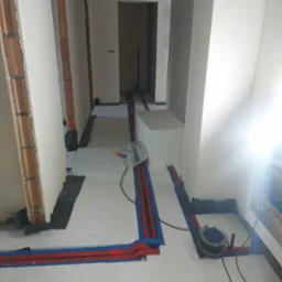 Wnętrze w trakcie instalacji ogrzewania podłogowego: rury grzewcze w kolorze czerwonym i niebieskim ułożone na białych płytach styropianowych, widoczne fragmenty ścian z surowej cegły...