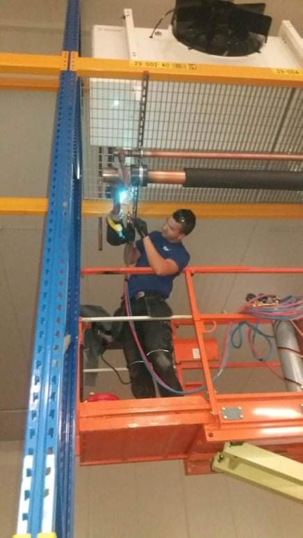 Instalator w niebieskiej koszulce spawa rury miedziane klimatyzacji na wysokości, stojąc na podnośniku koszowym w magazynie z regałami.