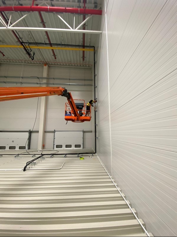Montaż instalacji w hali magazynowej z użyciem podnośnika koszowego; widoczne rury wentylacyjne pod sufitem i białe panele ścienne.