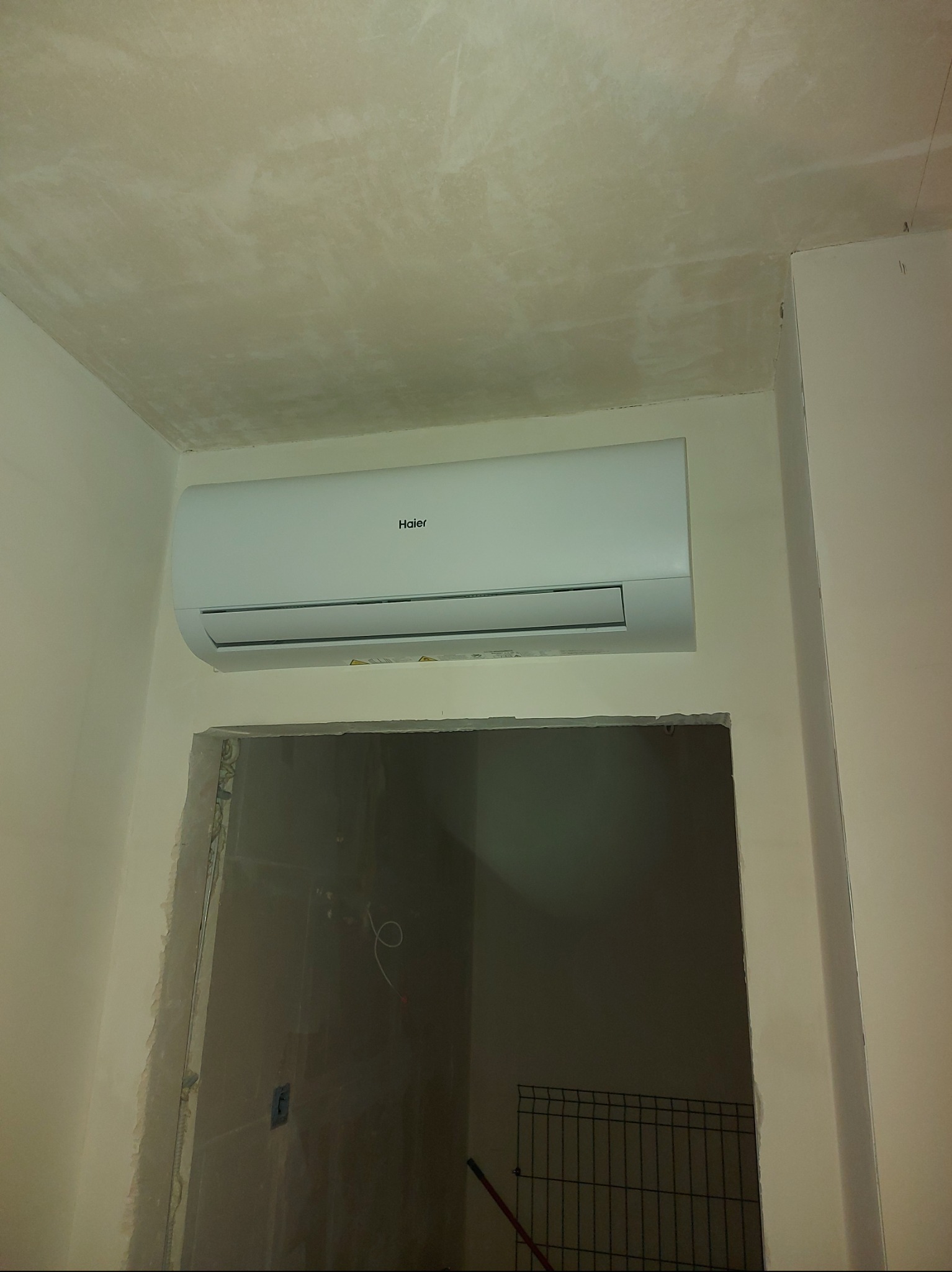 Biała klimatyzacja Haier zamontowana na ścianie nad otworem drzwiowym w trakcie remontu. Widoczne niedokończone wykończenia i surowy sufit.