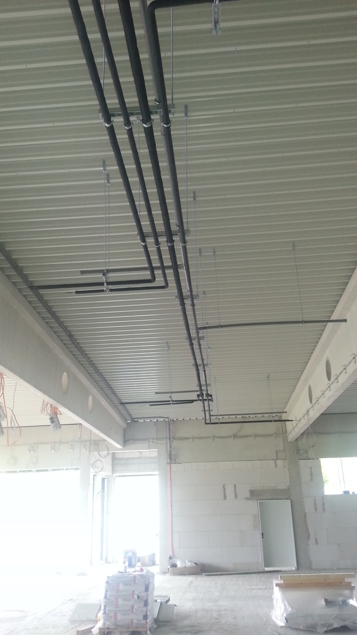 Montaż instalacji klimatyzacji: czarne rury pod sufitem z blachy trapezowej, mocowane na metalowych uchwytach w surowym wnętrzu budynku.