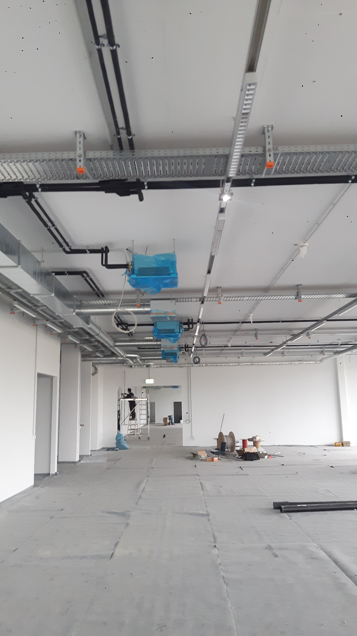 Montaż klimatyzacji w biurze: widok na sufit z instalacjami, klimatyzatory w niebieskiej folii ochronnej, pracownik na drabinie. Przestrzeń biurowa w trakcie wykańczania.