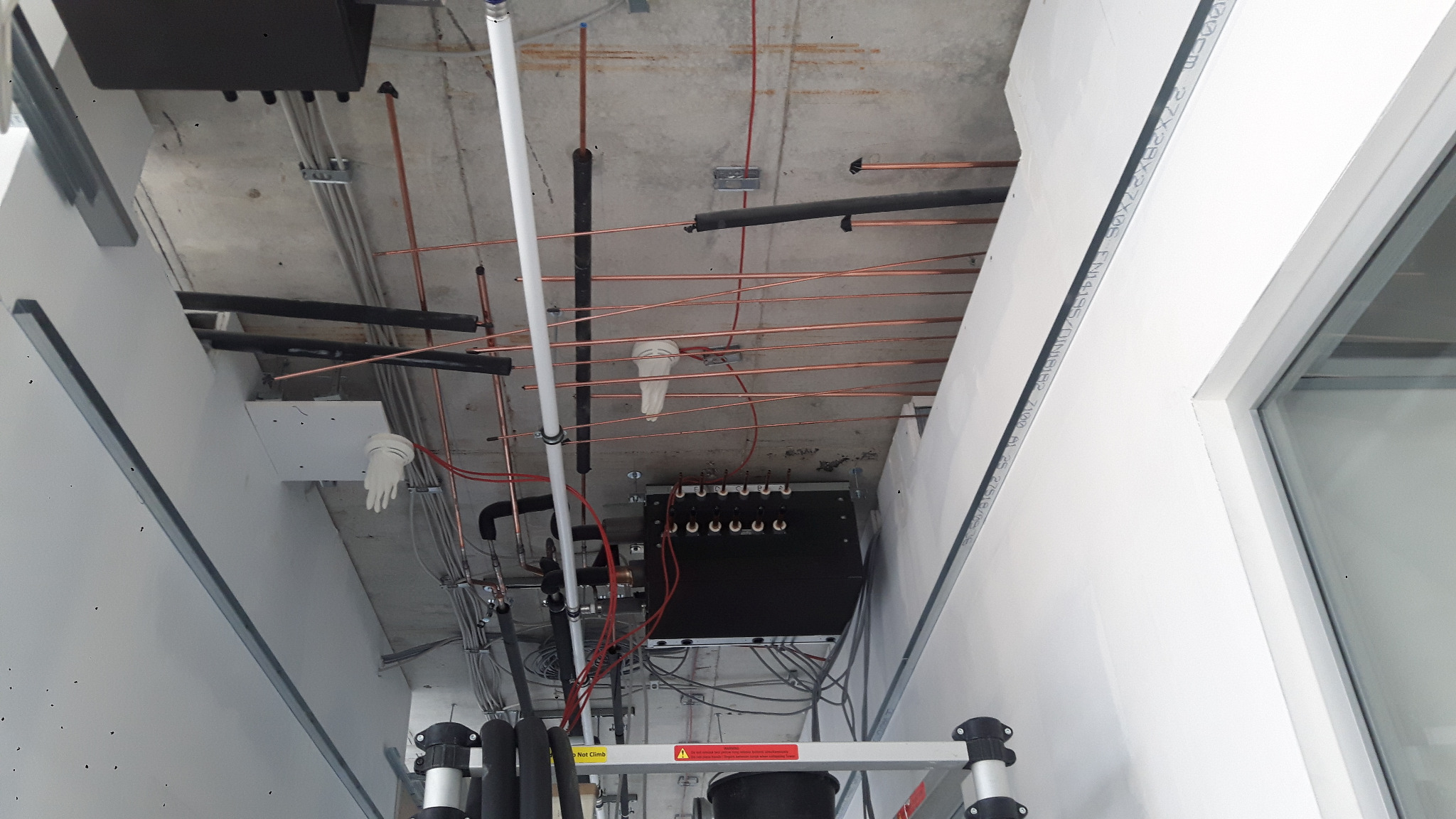 Montaż instalacji klimatyzacji: miedziane rury, czarne izolacje, białe przewody, rozdzielnica na metalowej konstrukcji, widok z drabiny, surowy betonowy sufit.