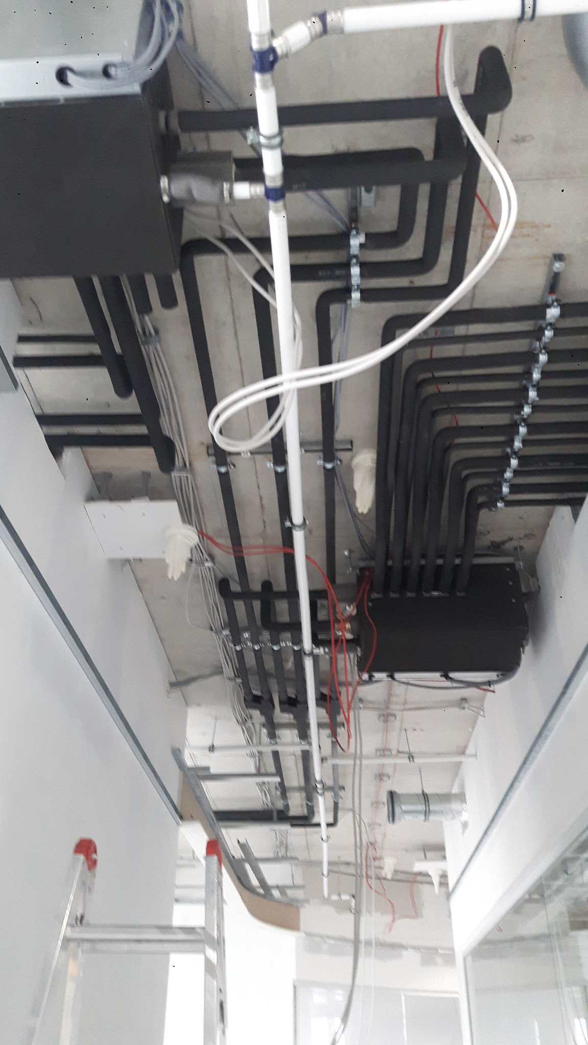 Instalacja klimatyzacji pod sufitem: czarne rury, białe przewody, elementy montażowe. Widoczna drabina i fragment okna. Ujęcie z dołu, podkreślające złożoność instalacji.