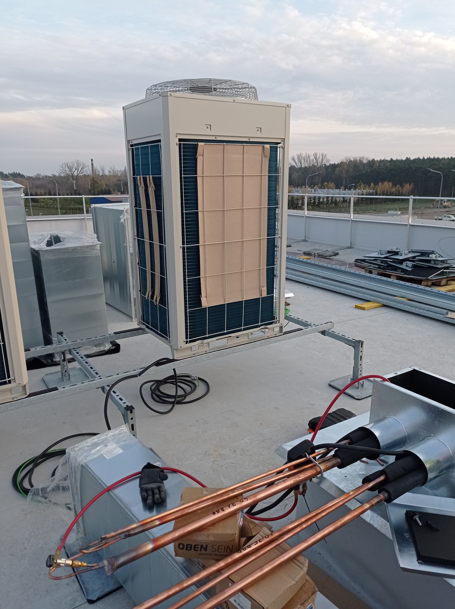 Montaż klimatyzacji na dachu budynku. Widoczne jednostki zewnętrzne, rury miedziane, elementy instalacyjne i narzędzia. Tło stanowi krajobraz miejski z drzewami.