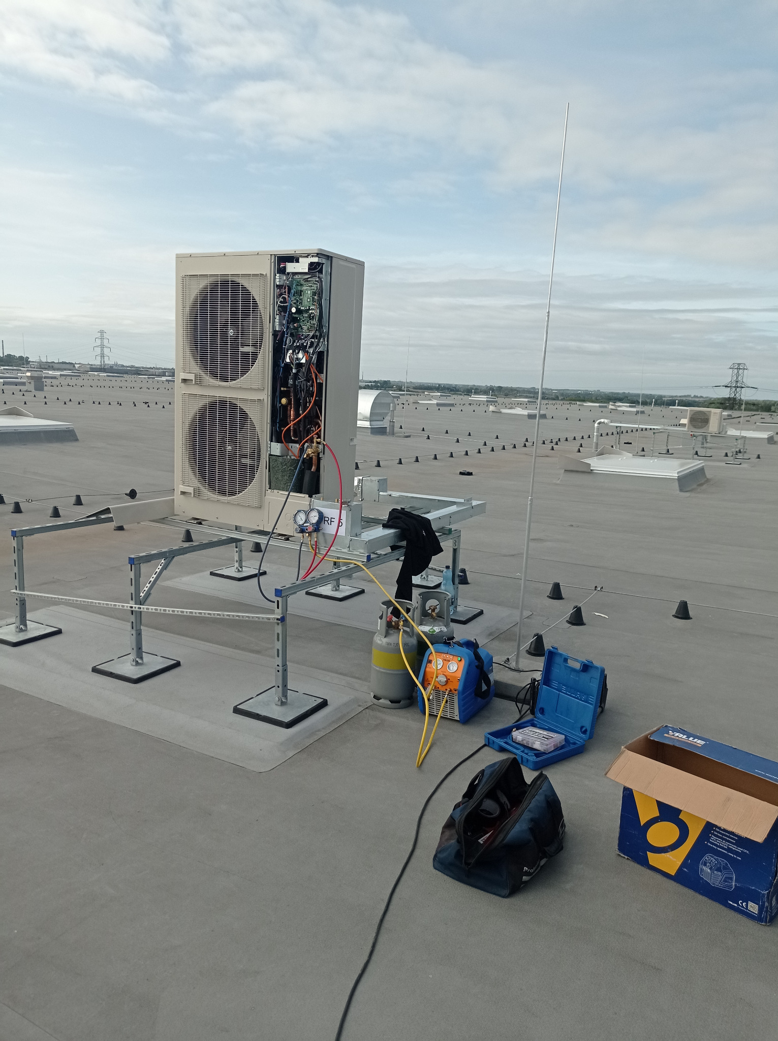 Serwis klimatyzacji na dachu budynku: otwarta obudowa jednostki zewnętrznej, podłączone manometry, butla z czynnikiem chłodniczym i narzędzia serwisowe.