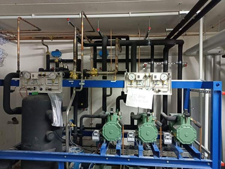 Kompleksowa instalacja klimatyzacyjna z trzema sprężarkami, manometrami i rurami miedzianymi, zamontowana na niebieskiej ramie. Widoczne przewody i zawory.