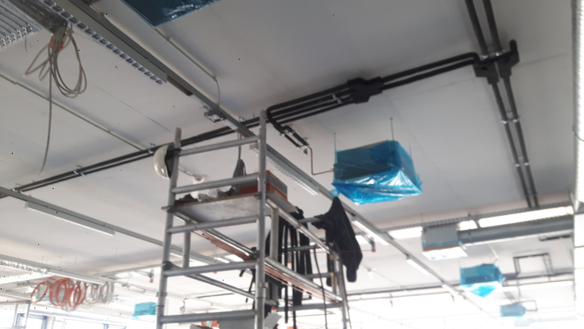Montaż instalacji klimatyzacji w biurze: rury, kable i jednostki wewnętrzne zabezpieczone folią, widoczne z poziomu rusztowania. Oświetlenie sufitowe.
