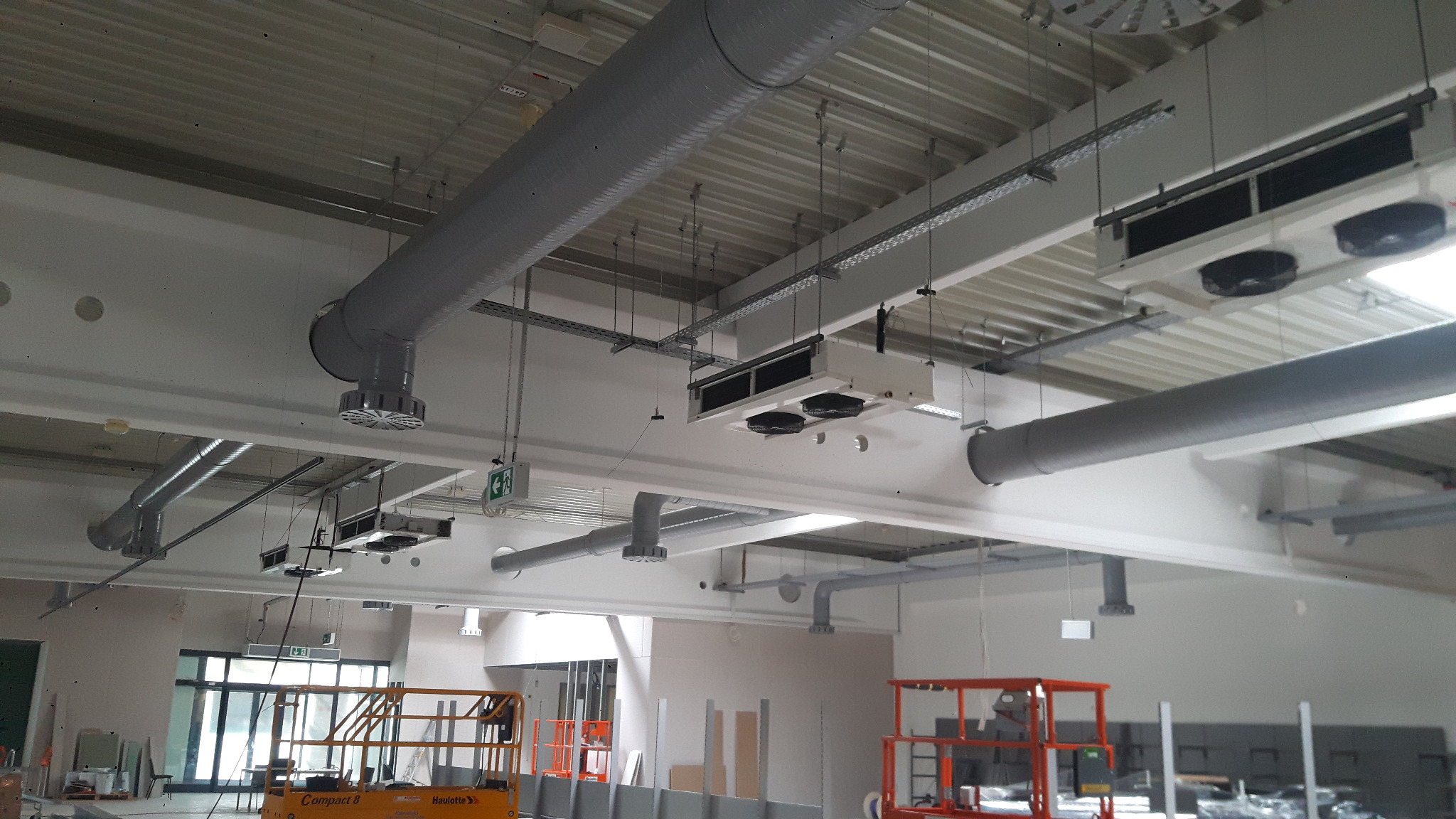 Szare rury wentylacyjne i białe klimatyzatory sufitowe w jasnym, przestronnym pomieszczeniu biurowym w trakcie instalacji. Widoczne podnośniki koszowe.