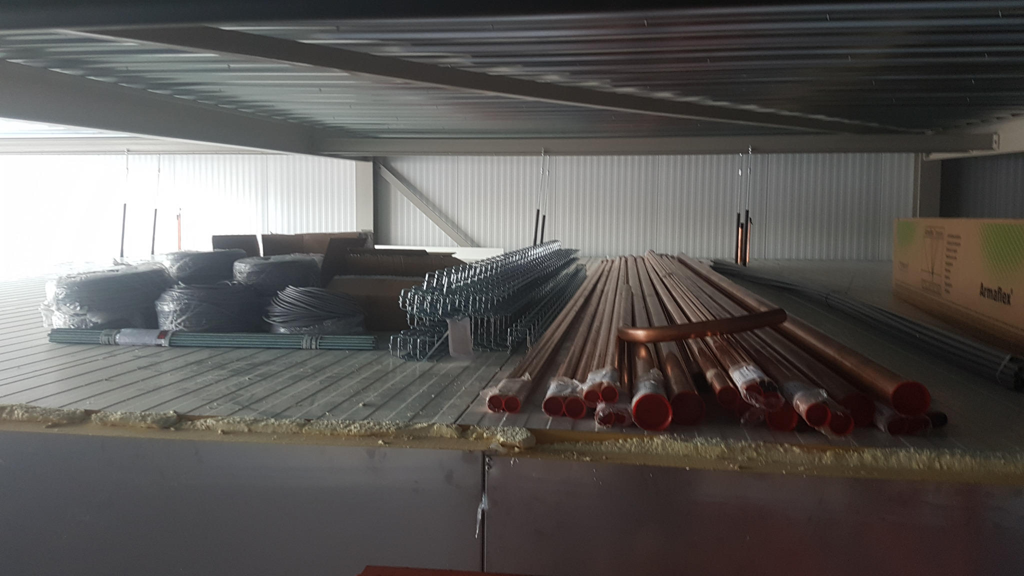 Miedziane rury, kable w izolacji i metalowe konstrukcje na podłodze w hali z blachy falistej, przygotowane do montażu instalacji. Widoczny karton Armaflex.