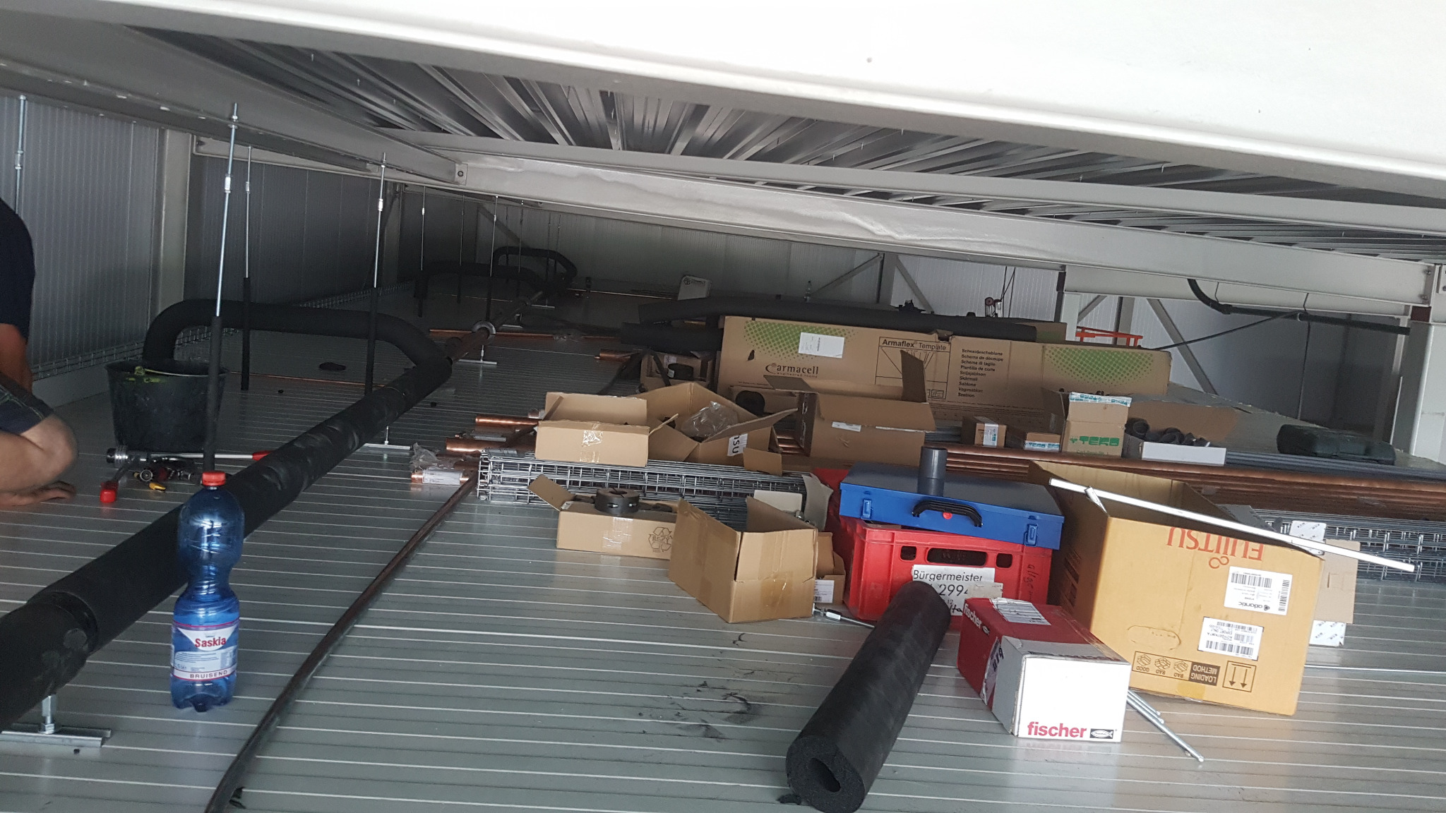 Montaż klimatyzacji w pomieszczeniu technicznym: widoczne rury, kable, narzędzia i materiały instalacyjne, butelka wody. Praca w toku, wnętrze hali.