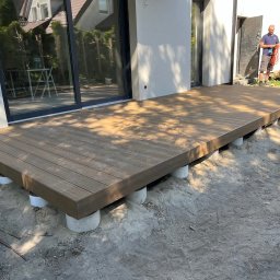 Vk-Bud - Nowoczesny taras z desek kompozytowych w odcieniach brązu, wsparty na betonowych słupkach. W tle widoczne okno tarasowe i sylwetka mężczyzny.