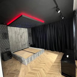 Vk-Bud - Nowoczesna sypialnia z szarym łóżkiem, parkietem w jodełkę, czarnym sufitem z oświetleniem LED i dekoracyjnymi zasłonami. Minimalistyczny design wnętrza.